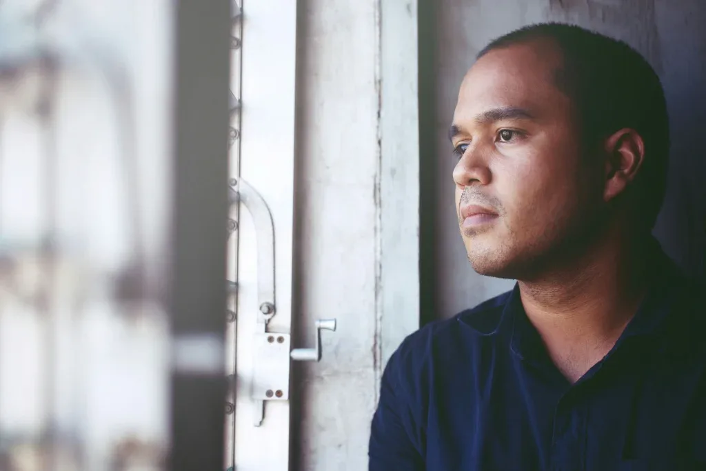 Man in dark shirt sitting by window, struggling with addiction and bipolar